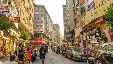 En travel handlegate i Istanbul med butikkskilt, parkerte biler og folk på fortauet
